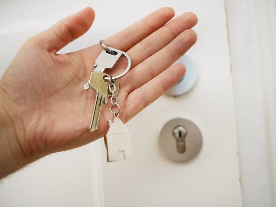 Offene Hand mit einem Schlüsselbund und Haus-Anhänger vor einer weißen Wohnungstür